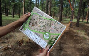 A person is holding up a map of the Flagstaff Barefoot Trail, with forest trees in the background.
