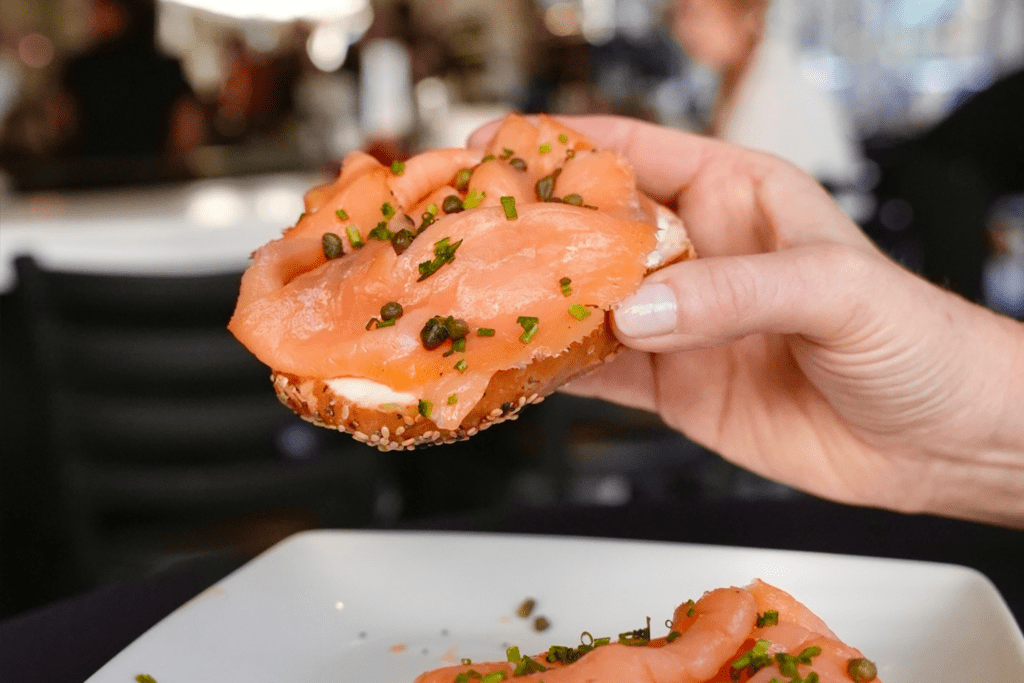 A person's hand holds a bagel with cream cheese, locks, chives and capers from Fortissimo Dueling Pianos.