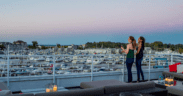 Two women stand next to a railing on the Marina Grand Rooftop Patio Lounge overlooking the harbor with sailing boats.