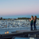 Two women stand next to a railing on the Marina Grand Rooftop Patio Lounge overlooking the harbor with sailing boats.
