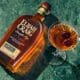 A bottle of Elijah Craig whiskey laying flat on a table next to a full martini glass full of whiskey with a double cherry garnish.