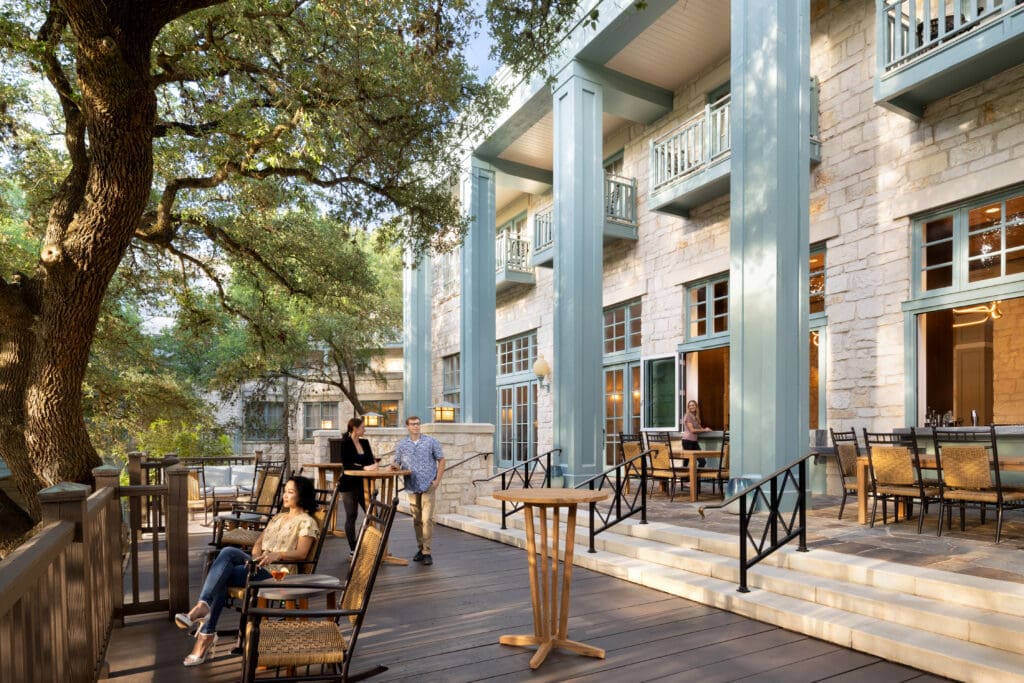 A person is sitting in a wood rocking chair on the patio of Hyatt Regency Hill Country Resort and Spa's Woodbine Bar close to nearby outdoor tables and seating.