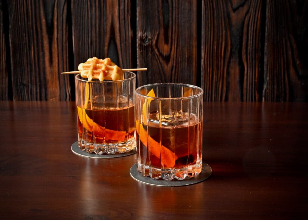 Two Berg Hospitality Group cocktails with orange peels and topped with a waffle sit on a wood bar.