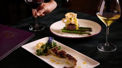 A person holds the stem of a red wine glass. Octopus and steak entrees are on the table with a glass of white wine.