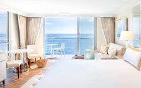A king-size bed, a couch, a table and two chairs with glass-front windows at Surf & Sand, Laguna Beach looking out to Laguna Beach.