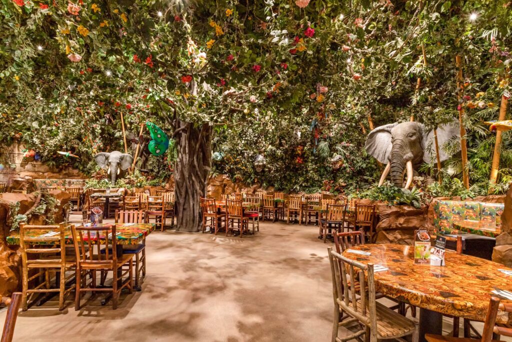 A huge tree with flowers with an elephant peeking out of the bushes and seating at the Rainforest Café.