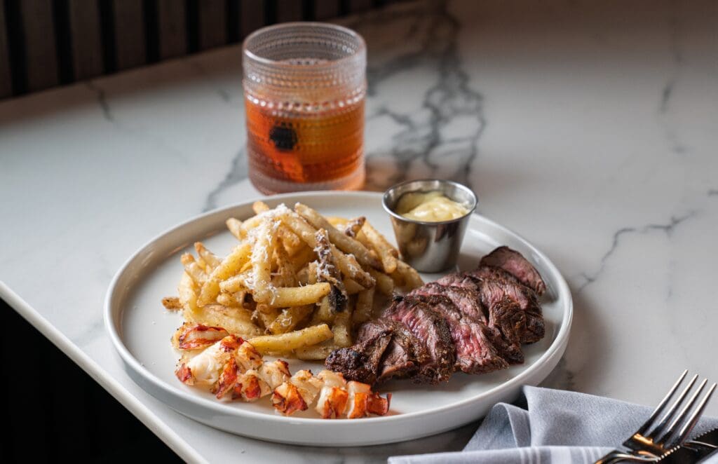A surf and turf plate with lobster tail, steak and truffle fries from MAVEN at Sawyer Yards with a cocktail on a table.