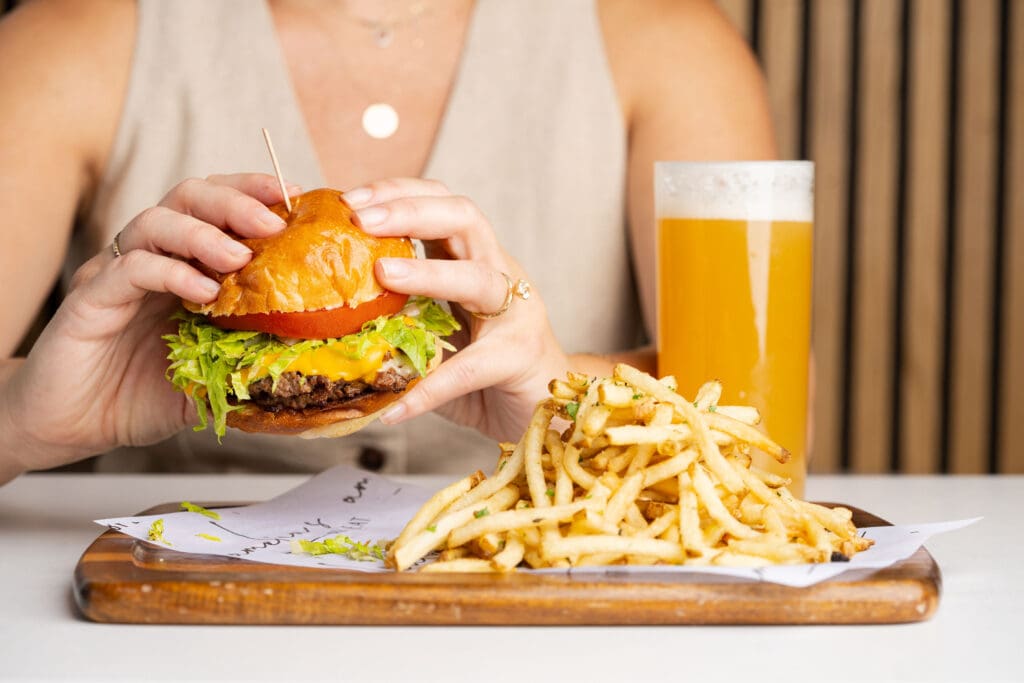 A woman holds a juicy burger in front of a cutting board with French Fries and a glass of beer.