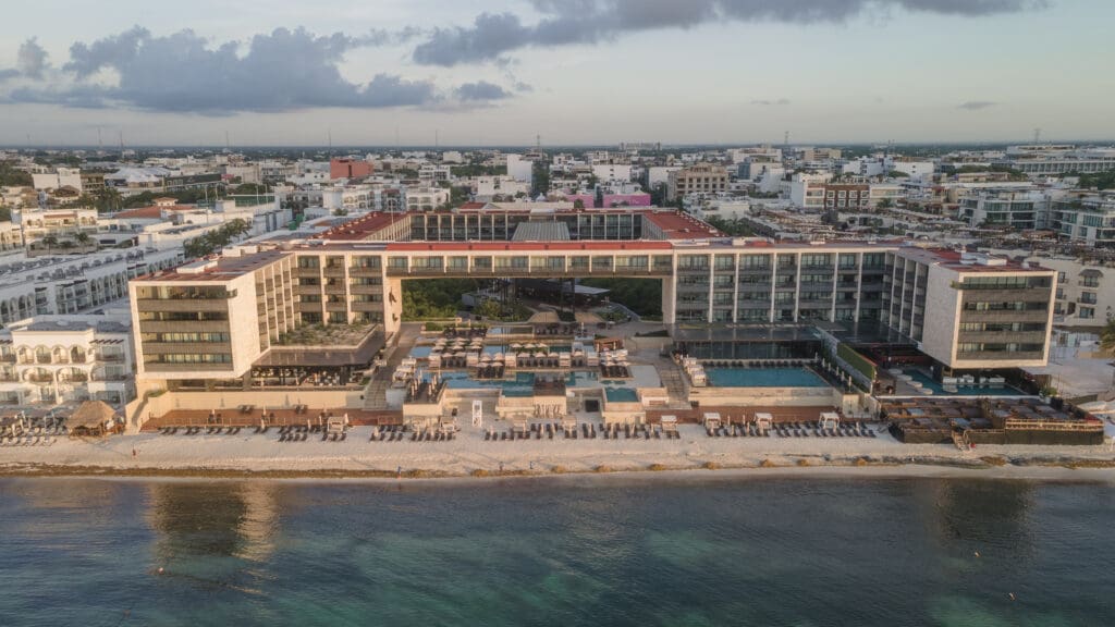 Devossion by Live Aqua adults-only hotel in Playa del Carmen showing the property and beachfront with the city in the background on a cloudy day.