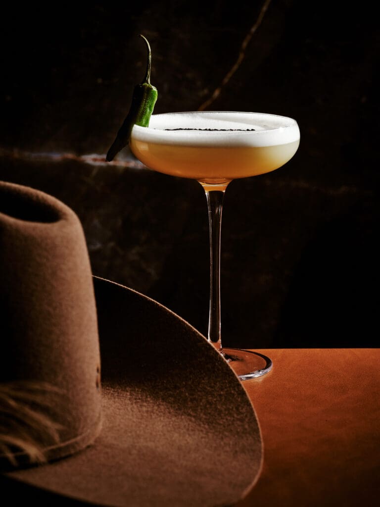 Kauboi's Shishito Sour signature cocktail adorned with a shishito pepper next to a Western hat at The REMI Scottsdale.