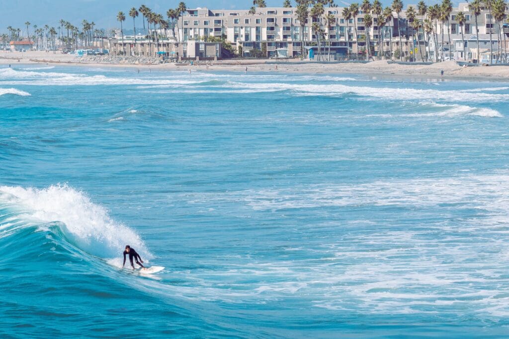 A surfer surfs a wave in Oceanside California with the beachfront in the distance.