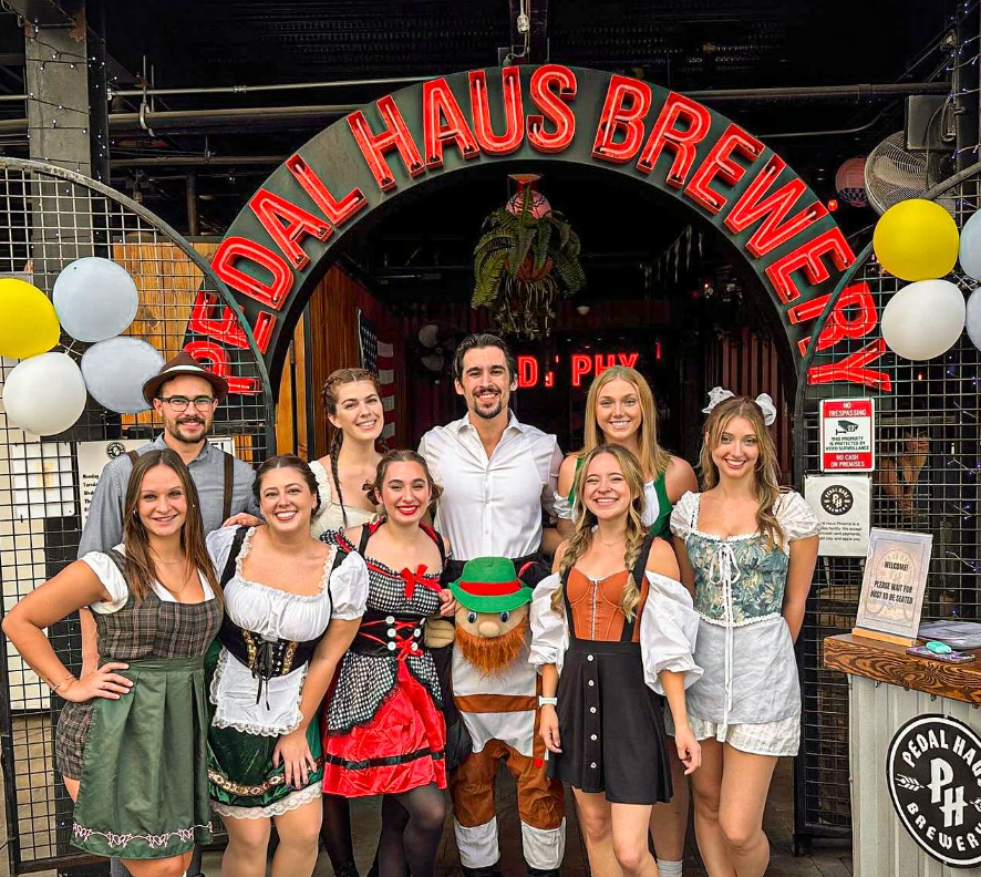 The Pedal Haus Brewery team in German attire at the Mesa location under a Pedal Haus neon sign for Oktoberfest.