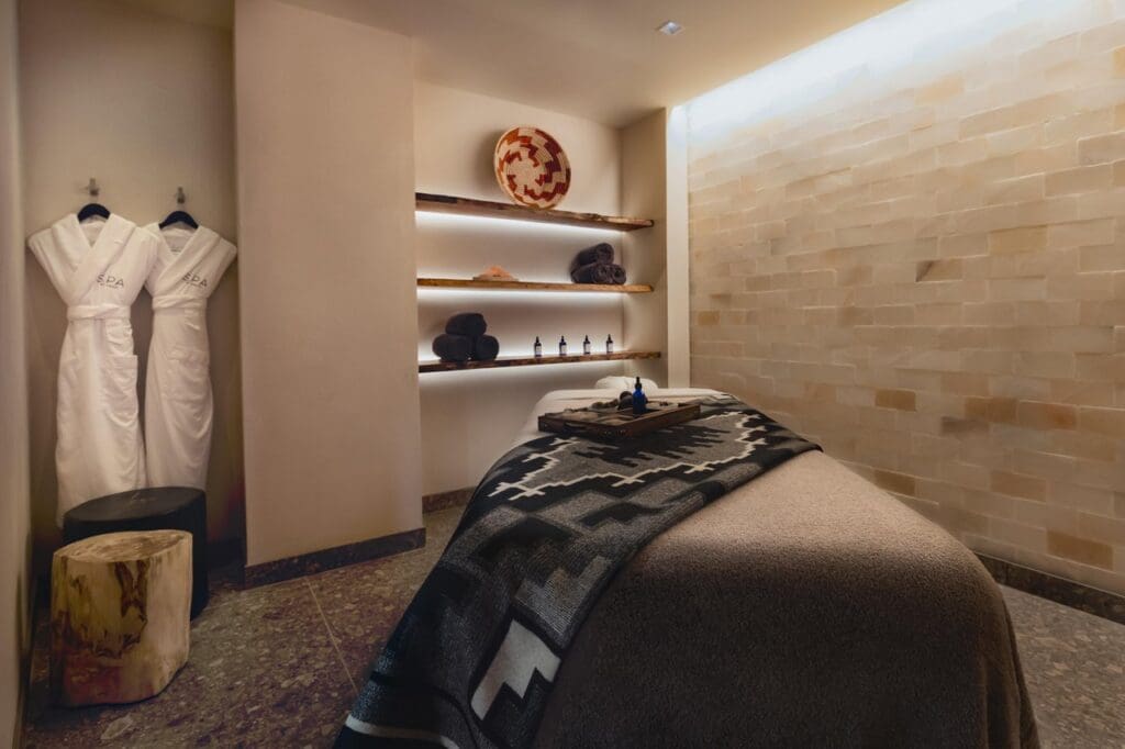 A massage room at The Spa at Chaco with two hanging robes, massage table with oils on a tray and native decor on shelves.