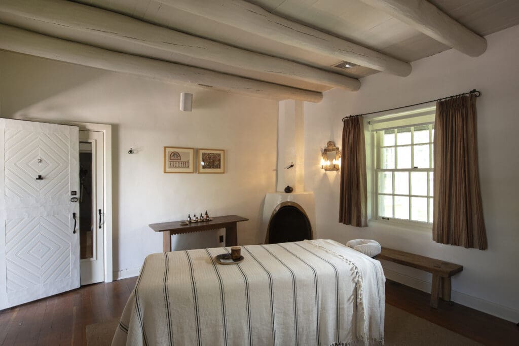 Massage room with adobe fireplace, massage table with tray with candle and sitting bench at Hacienda Spa at Los Poblanos.