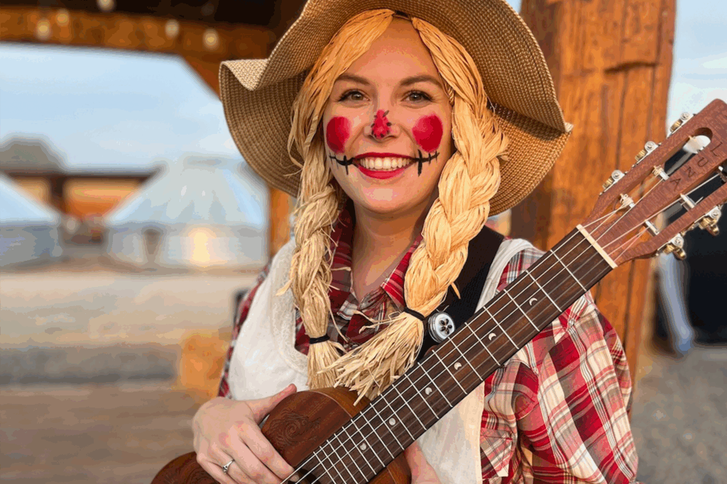 A woman dressed as a scarecrow with yellow straw braids, face paint and hat holds a guitar at Cornbelly's Corn Maze.