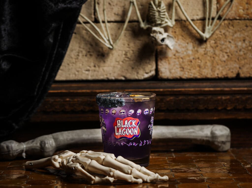 A Corpse Flower cocktail in a Black Lagoon cocktail glass, surrounded by a human hand skeleton and femur and skeletal upside down bat.
