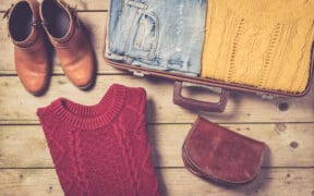 A pair of brown boots a red cable sweater, a pair of jeans and yellow cable sweater in a suitcase and a fall fashion purse.