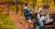 Three friends resting after hiking in forest, sitting on a collapsed trunk and drinking tea.