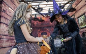 A little girl with curly hair and a cat cosume recieves candy in her bucket from a woman in a witch costume at Great Wolfe Lodge.