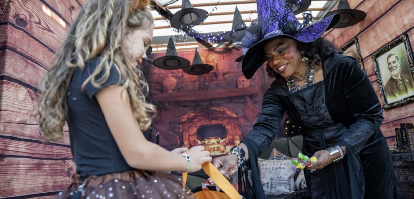 A little girl with curly hair and a cat cosume recieves candy in her bucket from a woman in a witch costume at Great Wolfe Lodge.