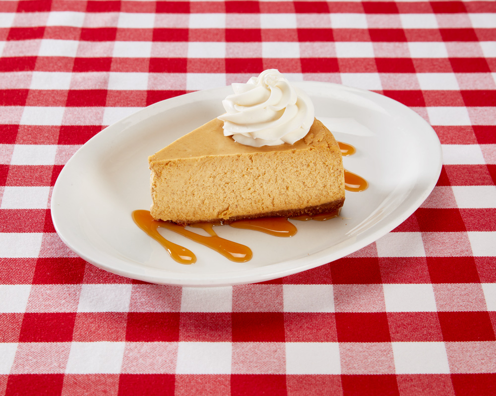 Pumpkin cheesecake topped with whipped cream on a layer of drizzled caramel plated on a table with a checkered tablecloth.