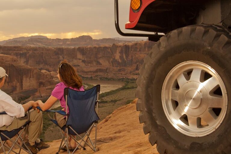 A couple sits side by side looking over a Moab national park cliff with a hummer behind them.