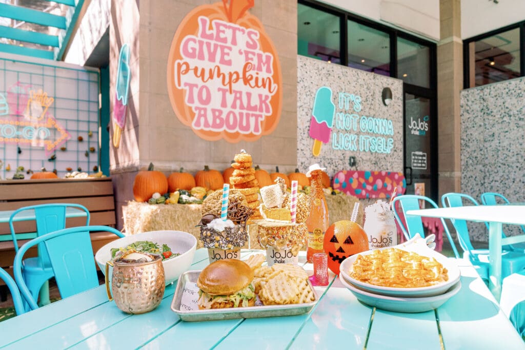 A Moscow Mule, hamburger and fries, two shakes and a waffle are on an outdoor table at fall-decorated JoJo's shakeBar.