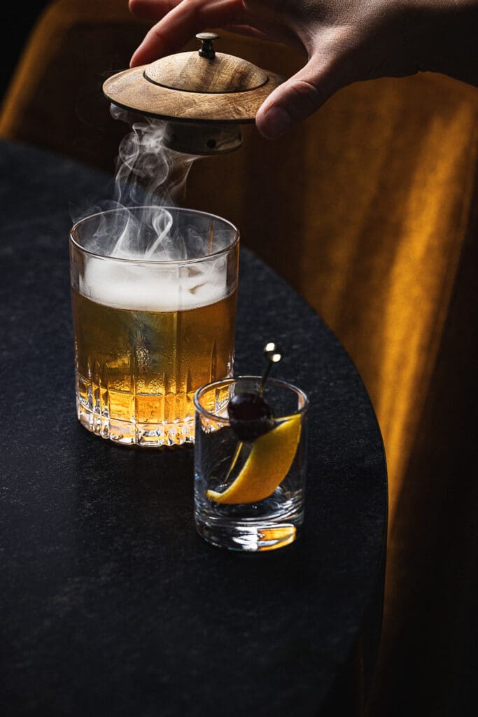 A person's hand lifting the lid off Sparrow's Ember Old Fashioned cocktail glass with a cherry and orange peel in a shot glass.