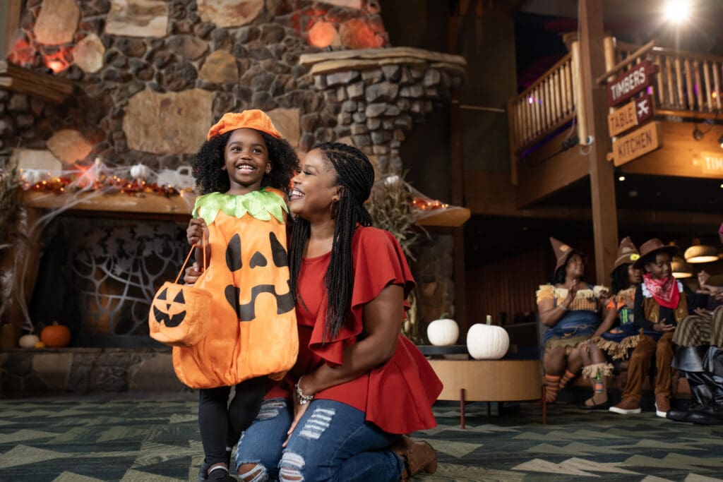 A mom kneels on the floor of the Great Wolf Lodge decorated for Halloween, hugging her daughter wearing a pumpkin costume, holding a pumpkin candy bag.