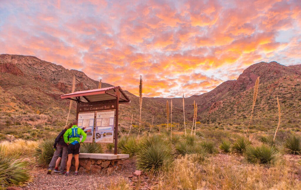 A small group of people stand by a trailhead at Franklin Mountains in El Paso with a pretty cloud sunrise above the mountains.