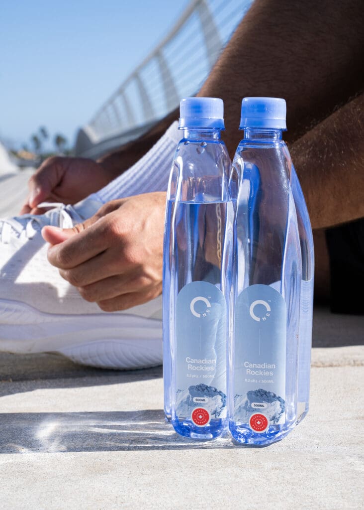 Two bottles of One Water spring water with a man tying his running shoes in the background. 