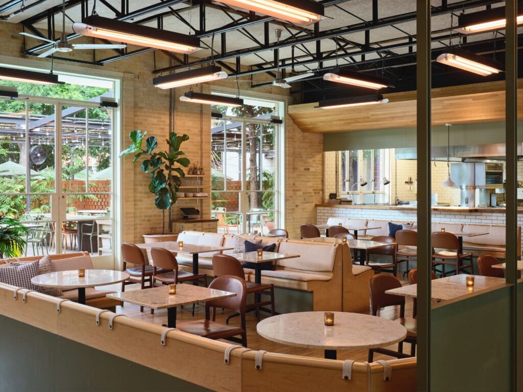 Tables with booths and chairs, each with a lit candle and an open chef's window at Carpenters Hall inside the Carpenter Hotel.