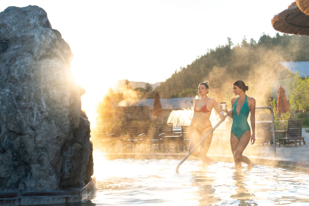 Two women in bathing suits, one holding a to-go coffee enter one of The Springs Resort's geothermal pools.