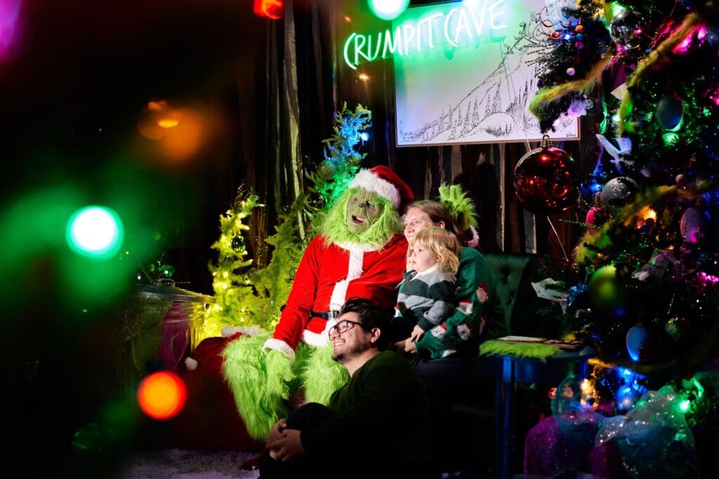 A mom, dad and toddler son sit with the Grinch dressed in Santa suit next to a Christmas tree in Denver's Dairy Block.