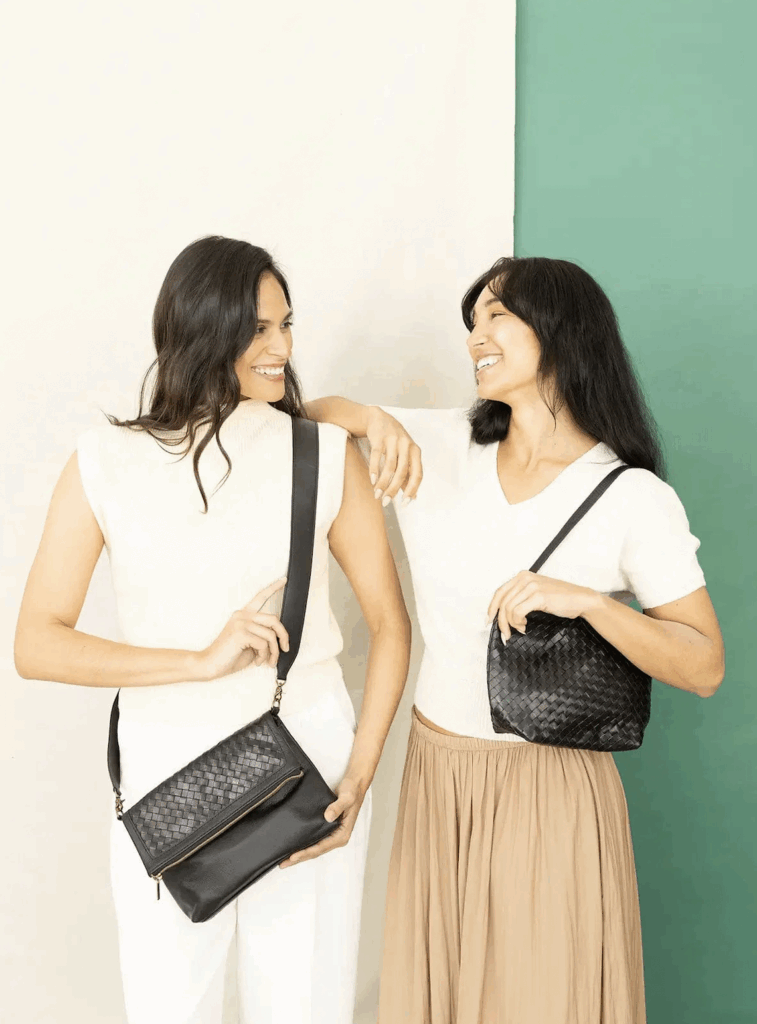 Two women smile at each other, both wearing Deux Mains, leather-woven purses.