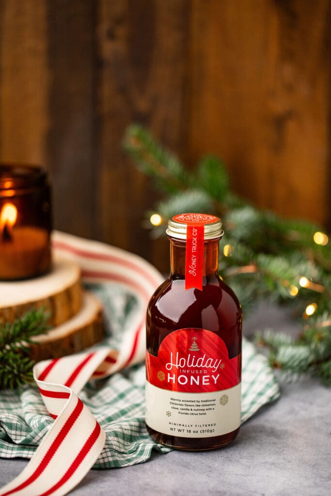 A bottle of Holiday Honey from The Honey Truck Co. is surrounded by holiday ribbon, a lit candle and greenery.