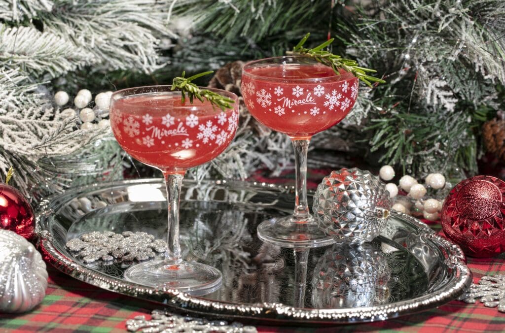 Two holiday beverages in snowflake cocktail stemware on a tray in front of a frosted Christmas tree and ornaments. 