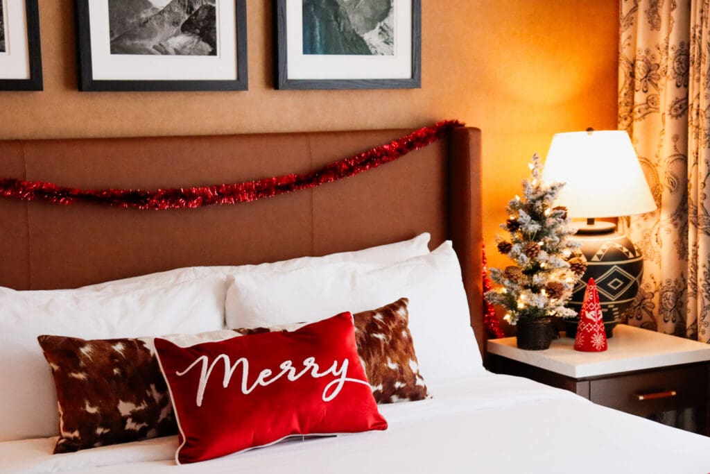 A bed at The Benson Hotel & Faculty Club in Aurora, Colorado with holiday pillows and a side table with decorative holiday trees. 