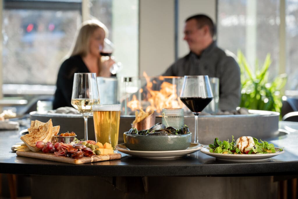 Beer, wine, a charcueterie board, oysters and a salad are on a table with a blurred couple next to a firepit in the background at the Lookout Bar at The Westin Riverfront Resort & Spa.
