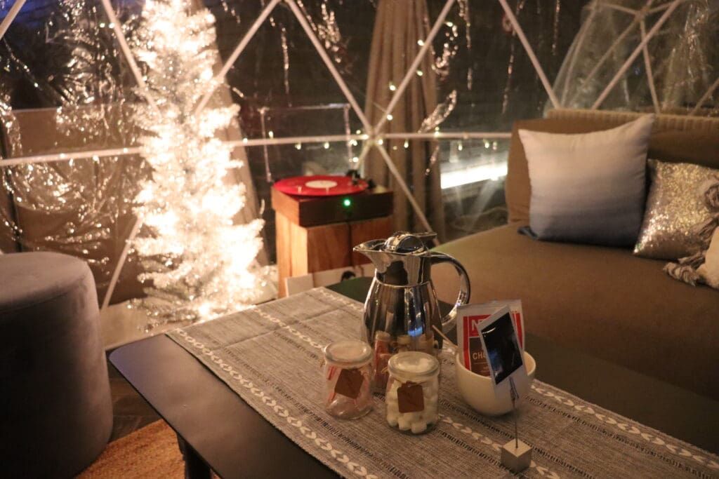 A decanter of hot chocolate, jar of marshmallows and candy canes on a table with couches inside a plastic snow globe at the Otis Hotel in Downtown Austin.