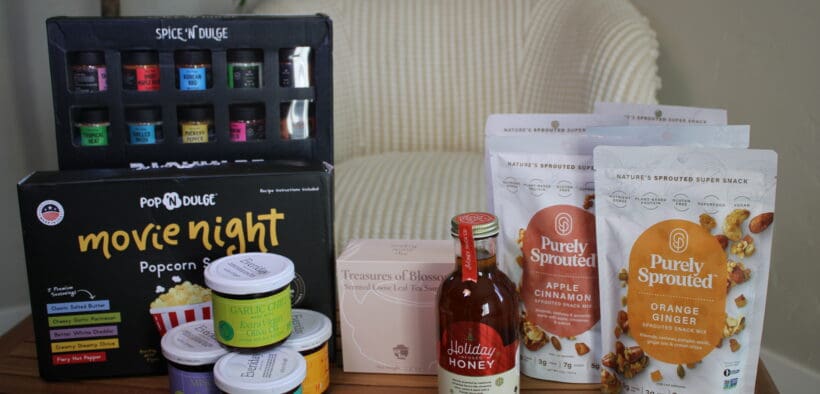 Pop 'n Dulge backyard spices, popcorn gift set, Eveiday chili sauces, a tea sampler set, Holiday Honey and Purely Sprouted snacks on a table.