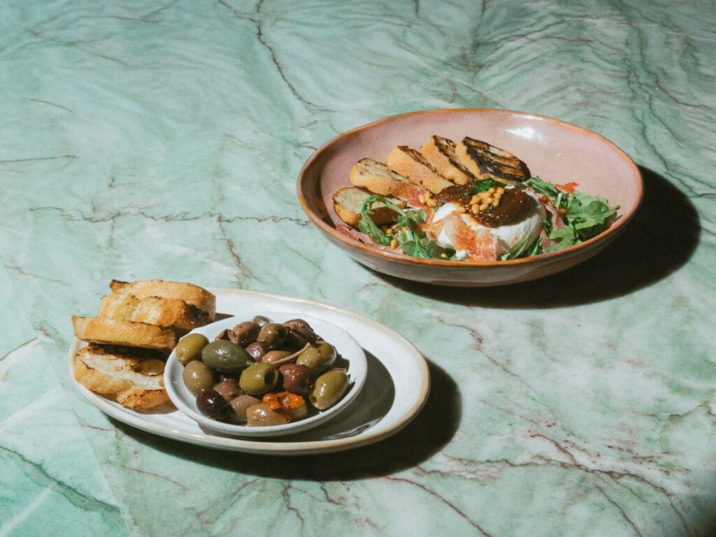A salad bowl with toasted bread slices and a bowl of mixed olives with bread slices at Estelle restaurant in Scottsdale.