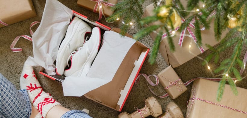 A person with pajama pants and Christmas socks opens a pressent with shoes inside and dumbbells nearby under a Christmas tree.
