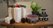 Several candles and a plant are on a table with a faux fur wrapped square present with a chair and plant in the background