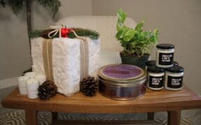 Several candles and a plant are on a table with a faux fur wrapped square present with a chair and plant in the background