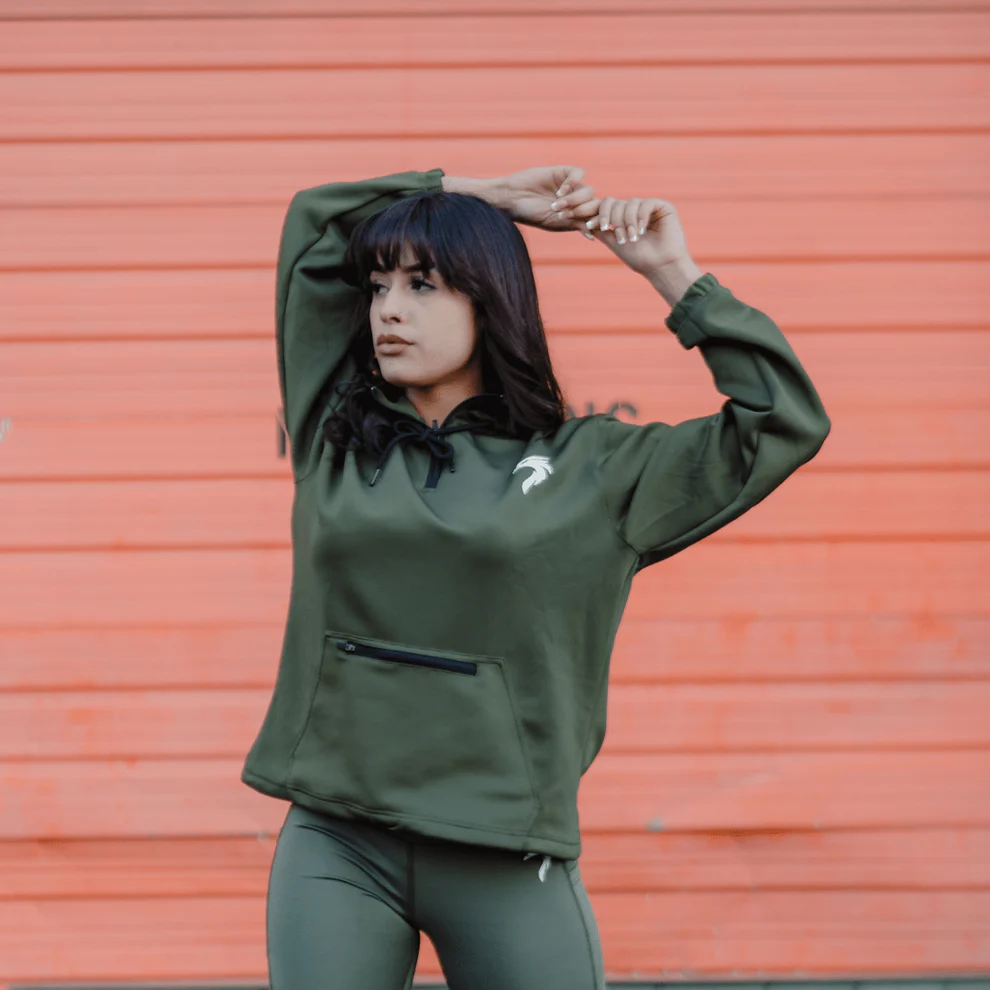 A woman stretches her arms while wearing an Army green Genesis Hoodie from Riseabove Apparel.