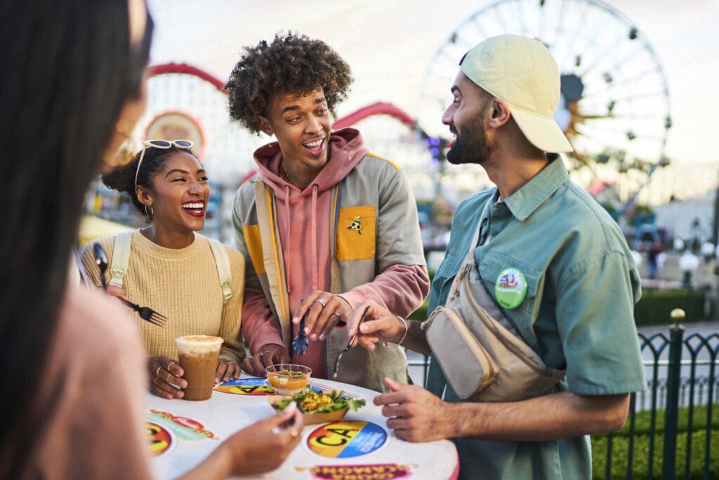 Two young people smile while speaking with a Disney cast member, enjoying a meal at the Disney Claifornia Adventure Food & Wine Festival.