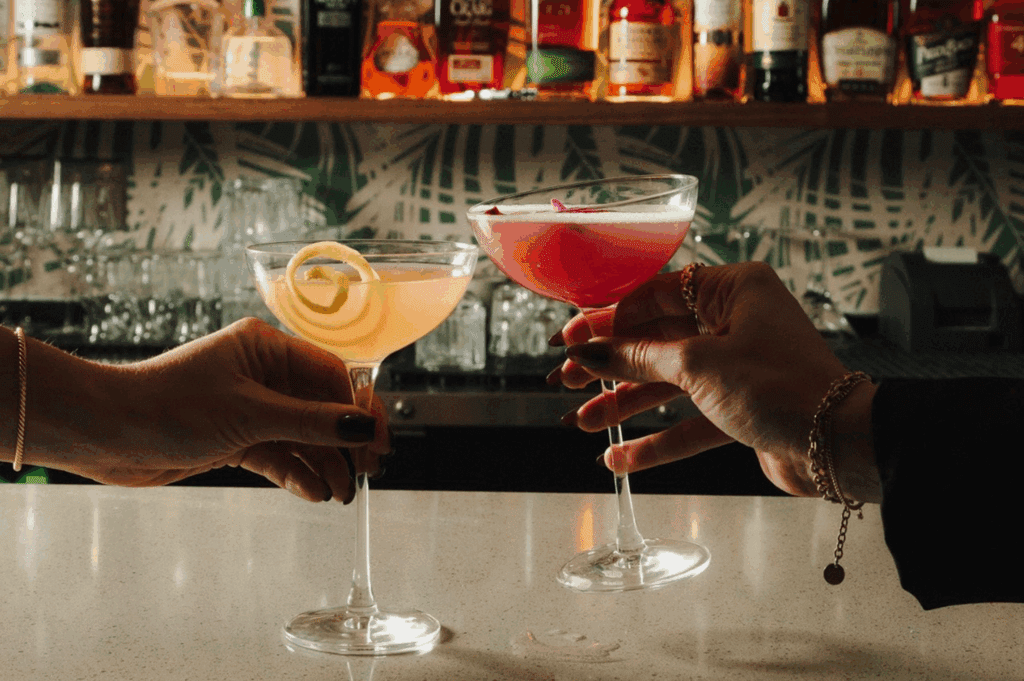 Two hands toast cocktail glasses with a row of liquor in the background at 40 Love.