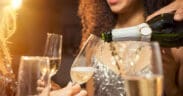 A hand pours champagne into flutes with a woman in a cocktail dress in the background on New Year's Eve.