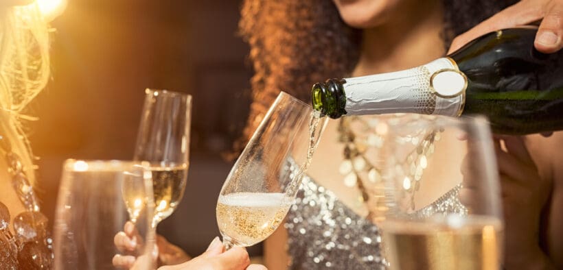A hand pours champagne into flutes with a woman in a cocktail dress in the background on New Year's Eve.